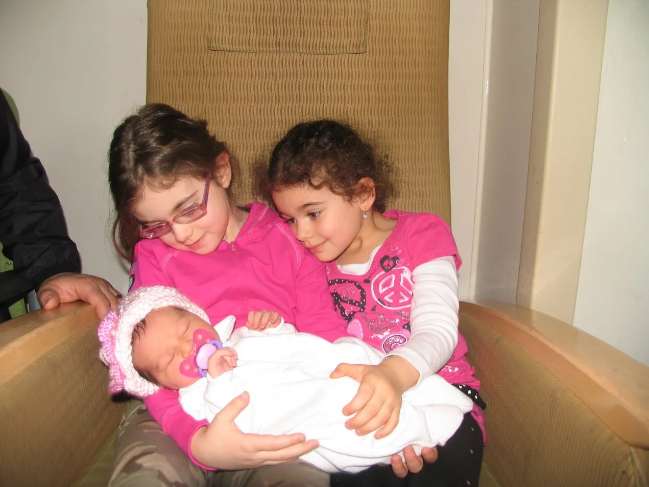 Two big sisters in pink holding their newborn baby sister in a pink knit hat