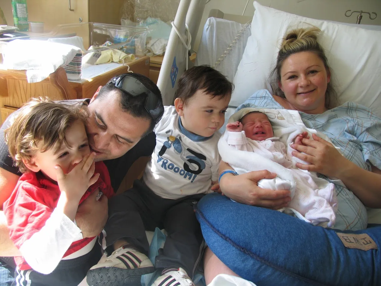 Mom in a hospital bed holding her newborn baby girl while dad and two young brothers gather close