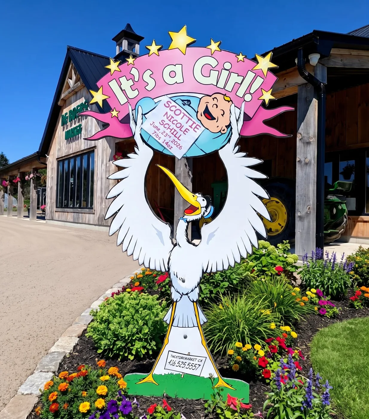 Pink It’s a Girl lawn stork sign for Scottie Nicole in a flower garden in front of a wooden barn market