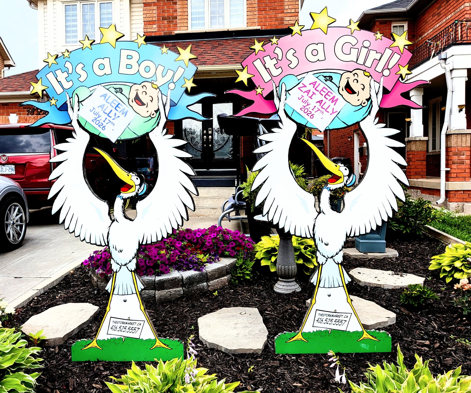Blue and pink twin lawn stork signs for Aleem Ally and Aleem Zai Ally in a flower bed — The Stork Market, serving Brampton and the GTA