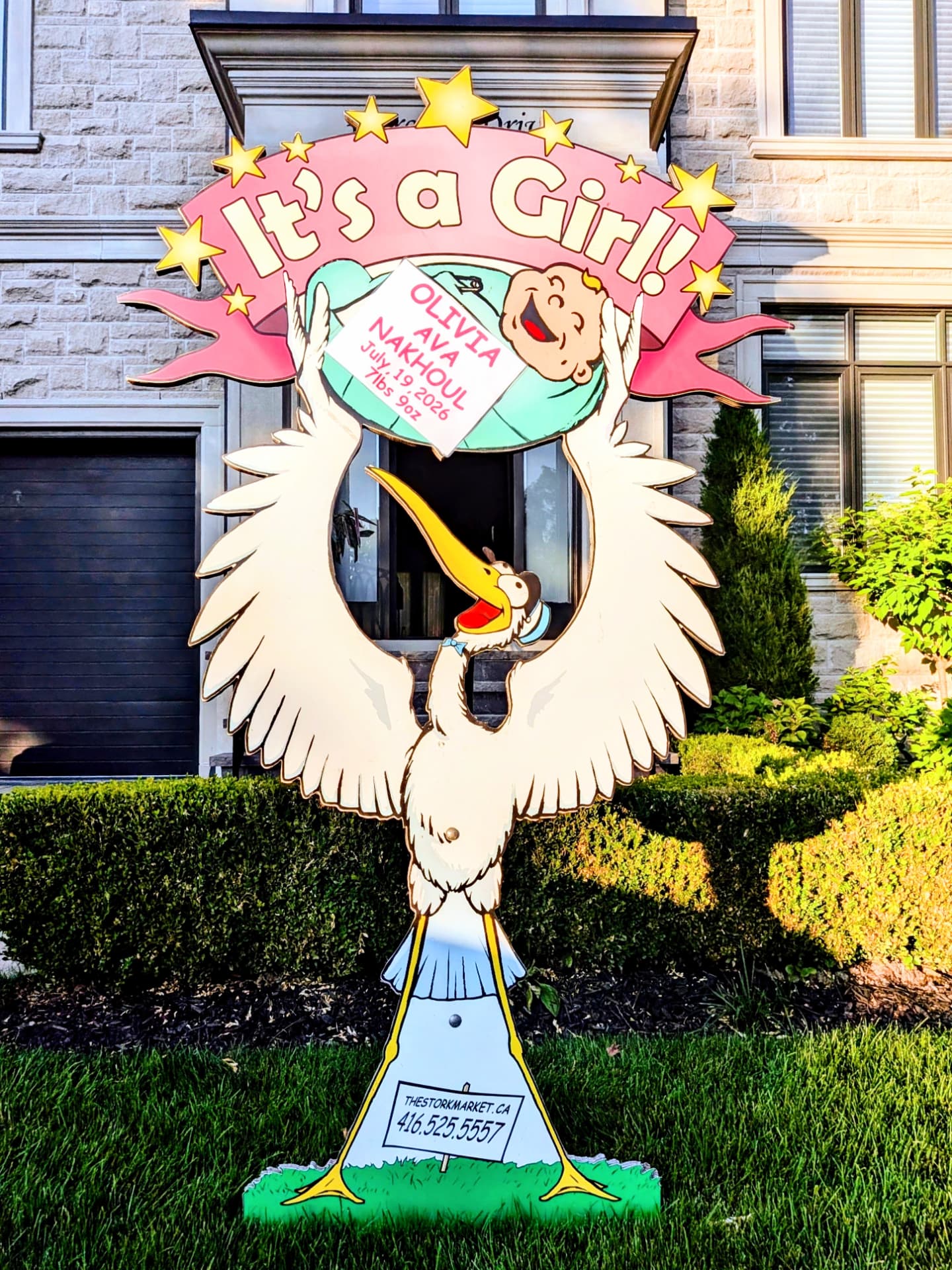 Pink Olivia Ava Nakhoul lawn stork sign illuminated at dusk by a house entrance — The Stork Market, serving Brampton and the GTA